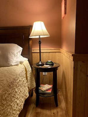 Una cama con edredón blanco y almohadas, una mesita de noche con lámpara, reloj y revista.