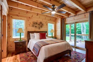 A wooden cabin bedroom with a bed, two nightstands, a ceiling fan, and large windows.