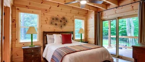 Un dormitorio de cabaña de madera con una cama, dos mesitas de noche, un ventilador de techo y ventanas amplias.