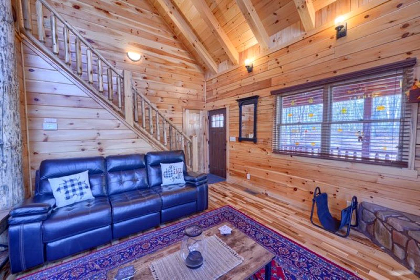 L’intérieur d’un chalet en bois, avec un escalier, un canapé en cuir noir, un tapis à motifs et une fenêtre avec des stores.