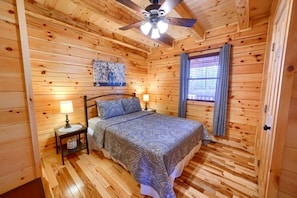 A wooden cabin bedroom with a bed, two bedside tables with lamps, a ceiling fan, and a window with curtains.