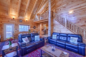 A log cabin interior with a living room featuring two leather sofas, a dining area, and a staircase leading to an upper level.