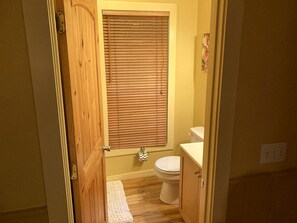 A bathroom with a wooden door, a toilet, a window with blinds, and a small rug on the floor. - Rustic Cabin on 28-Acre Property in the Foothills of the Blue Ridge Mountains (Elkin)