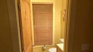 A bathroom with a wooden door, a toilet, a window with blinds, and a small rug on the floor. - Rustic Cabin on 28-Acre Property in the Foothills of the Blue Ridge Mountains (Elkin)