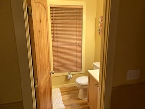 A bathroom with a wooden door, a toilet, a window with blinds, and a small rug on the floor.