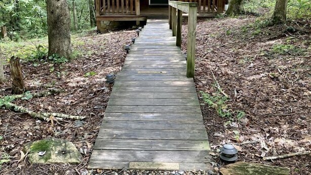 A wooden cabin with a porch, surrounded by trees and a wooden pathway leading to it. - Rustic Cabin on 28-Acre Property in the Foothills of the Blue Ridge Mountains (Elkin)