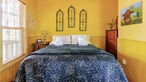 A hotel room with a bed, a nightstand, a lamp, a dresser, and a painting on the wall. - Rustic Cabin on 28-Acre Property in the Foothills of the Blue Ridge Mountains (Elkin)