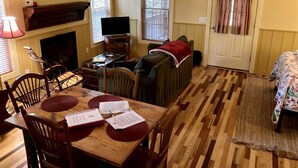 A cozy living room with a fireplace, a television, a dining table with chairs, a sofa, and a door with curtains. - Rustic Cabin on 28-Acre Property in the Foothills of the Blue Ridge Mountains (Elkin)