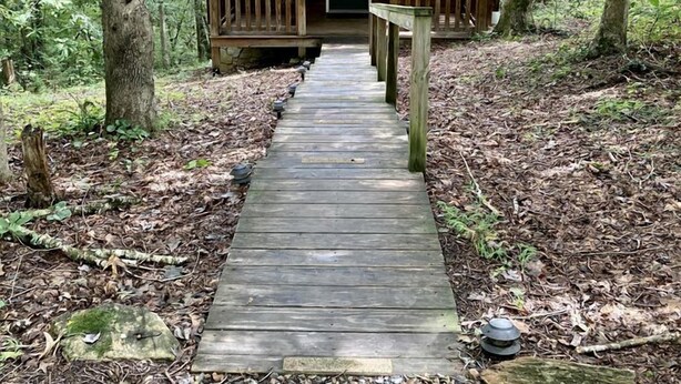 A wooden cabin with a porch, surrounded by trees and a wooden pathway leading to it. - Rustic Cabin on 28-Acre Property in the Foothills of the Blue Ridge Mountains (Elkin)