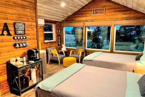 A cozy cabin-style room with wooden walls, two beds, a coffee station, and large windows offering a view of the outdoors.