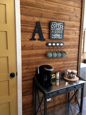 Pared de madera con un letrero alusivo al café, una cafetera y tazas sobre una mesa.