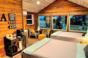 Una habitació acollidora d'estil cabana, amb parets de fusta, dos llits, racó de cafè i grans finestres amb vista a l'exterior.