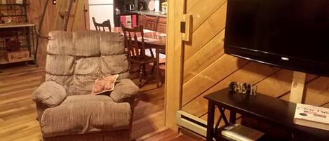 Un salon chaleureux avec un grand téléviseur à écran plat, un fauteuil confortable et une table à manger en bois.