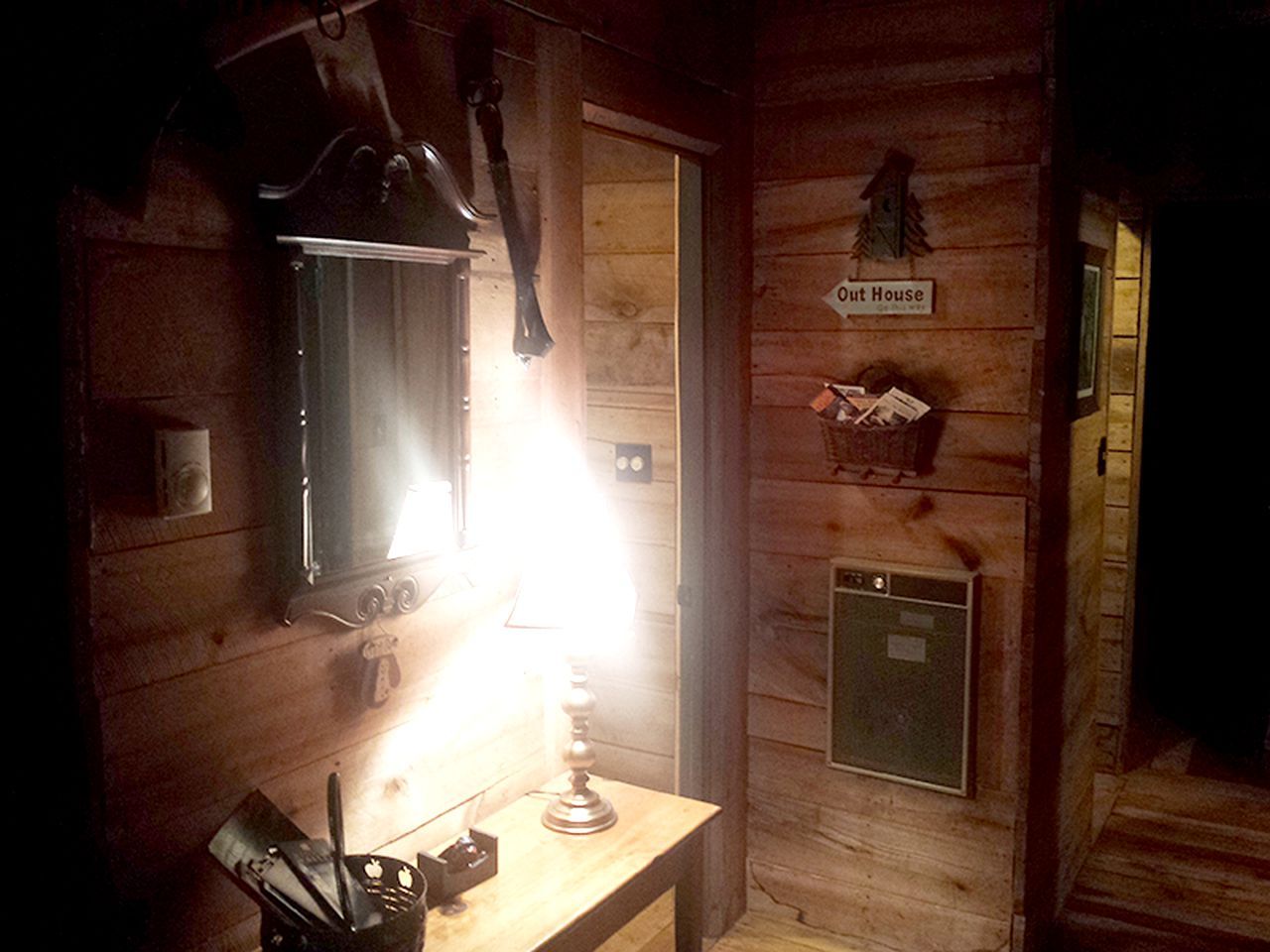A wooden cabin interior with a desk, a lamp, a mirror, a small cabinet, and a sign reading 'Out House.'