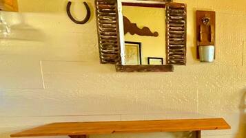 A room with a wooden table and two black bar stools, a mirror with a decorative frame, and a wall-mounted light fixture.
