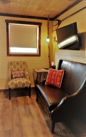 A room with a wooden ceiling, a sofa, a chair, a small table, and a TV mounted on the wall.