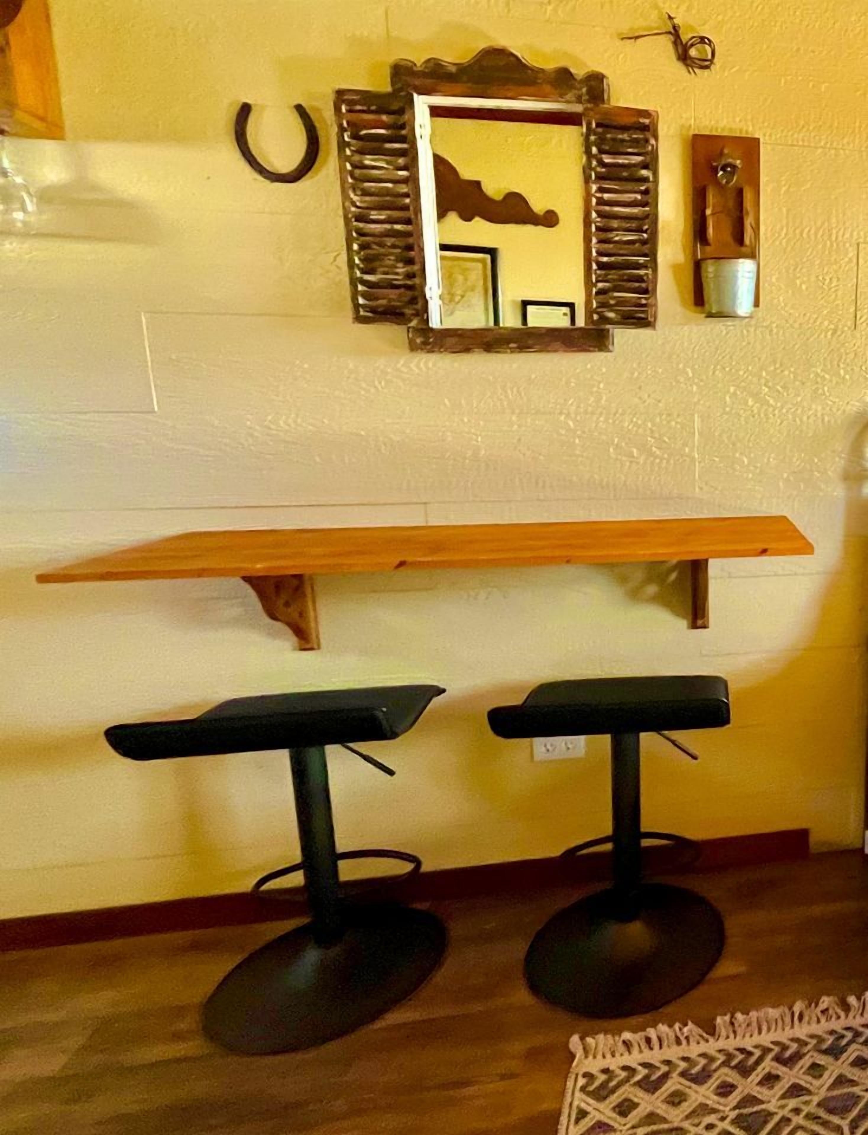 A room with a wooden table and two black bar stools, a mirror with a decorative frame, and a wall-mounted light fixture.