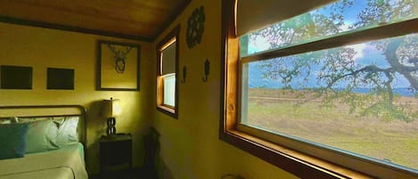 Ein Zimmer mit einem Bett, einem Nachttisch, einem Fenster mit Blick auf Bäume und einer Wanduhr.