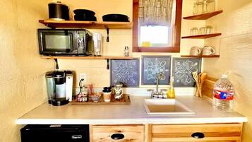 A compact kitchen with a microwave, coffee maker, and a small refrigerator. There are wooden cabinets and a window with a curtain.