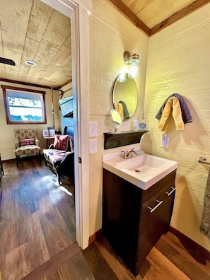 A compact room with a wooden ceiling, a small sink with a mirror, a chair, a sofa, and a window with a view.
