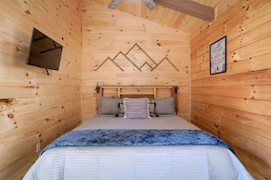 A wooden cabin room with a bed, a mounted TV, and a framed picture on the wall.