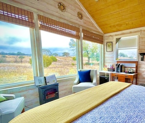 Une chambre avec un lit, un bureau et une chaise, de grandes fenêtres donnant sur la campagne et un plafond en bois.