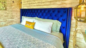 Een slaapkamer met een blauw gestoffeerd hoofdeinde, een bed met beddengoed met een patroon, een wandlamp en een rustieke houten wand.