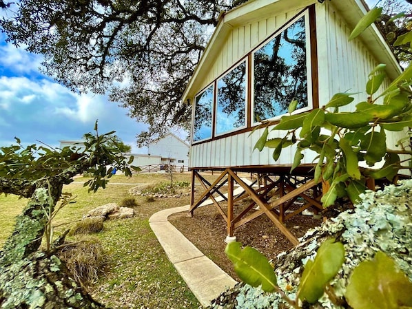 A wooden cabin with large windows, surrounded by a garden with various plants and a pathway leading to it. - Charming Texas Tree House with Tranquil Views Ideal for Glamping near Austin (Dripping Springs)
