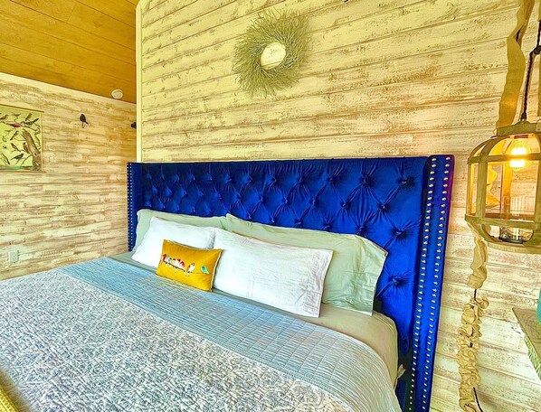 A bedroom with a tufted blue headboard, a bed with patterned bedding, a wall-mounted light fixture, and a rustic wooden wall.