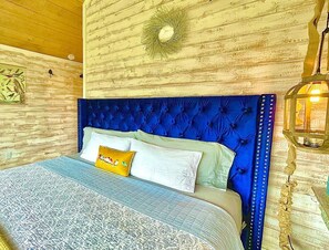 Een slaapkamer met een blauw gestoffeerd hoofdeinde, een bed met beddengoed met een patroon, een wandlamp en een rustieke houten wand.