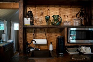 A kitchen with a wooden shelf, a kettle, a coffee maker, a microwave, and a sink.