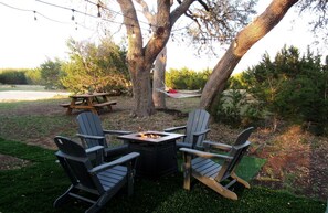 An outdoor seating area with Adirondack chairs and a square table, surrounded by trees and a picnic table.