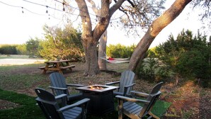 An outdoor seating area with Adirondack chairs and a square table, surrounded by trees and a picnic table. - Cozy Texas Hill Country Rental for Romantic Getaways in Texas (Dripping Springs)