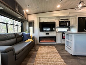 A modern RV interior with a sofa, fireplace, kitchen area, and large windows.