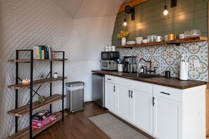 Cocina compacta con muebles blancos, encimera de madera y microondas. Se observan estantes con libros y objetos decorativos, además de un cubo de basura.