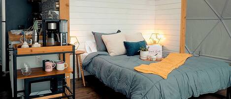 A compact bedroom with a bed, bedside table, and a small desk with a coffee machine.