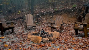 A forest setting with a fire pit and four Adirondack chairs.