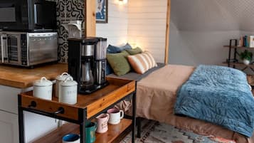A compact room with a kitchenette, a bed with a blue blanket, and a patterned rug.
