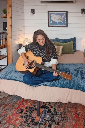 En person sitter på en säng och spelar gitarr i ett rum med en karta på väggen och en mönstrad matta.