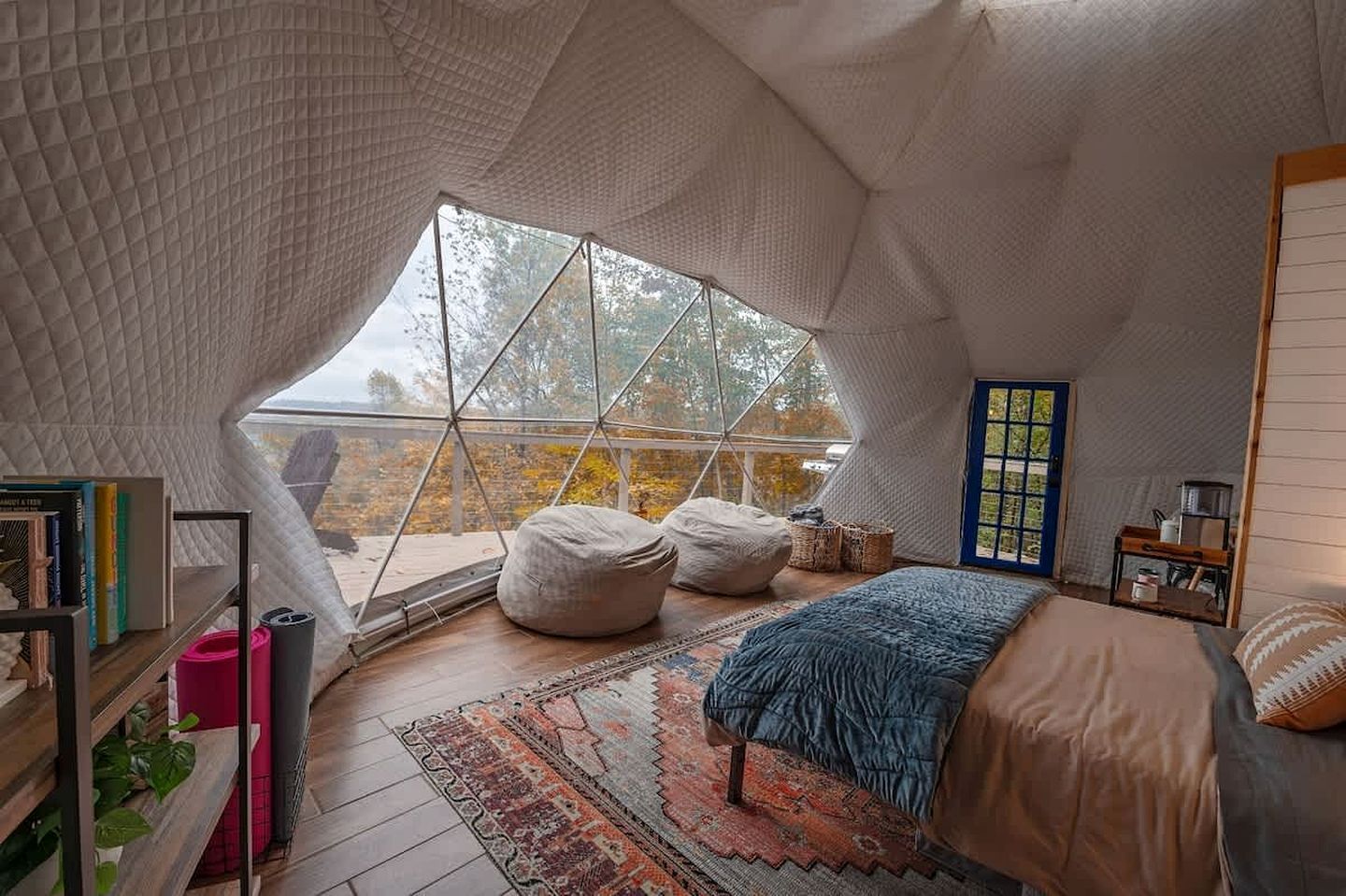 Um quarto aconchegante, com formato de tenda, que possui uma janela grande, uma cama, um tapete e uma estante de livros.