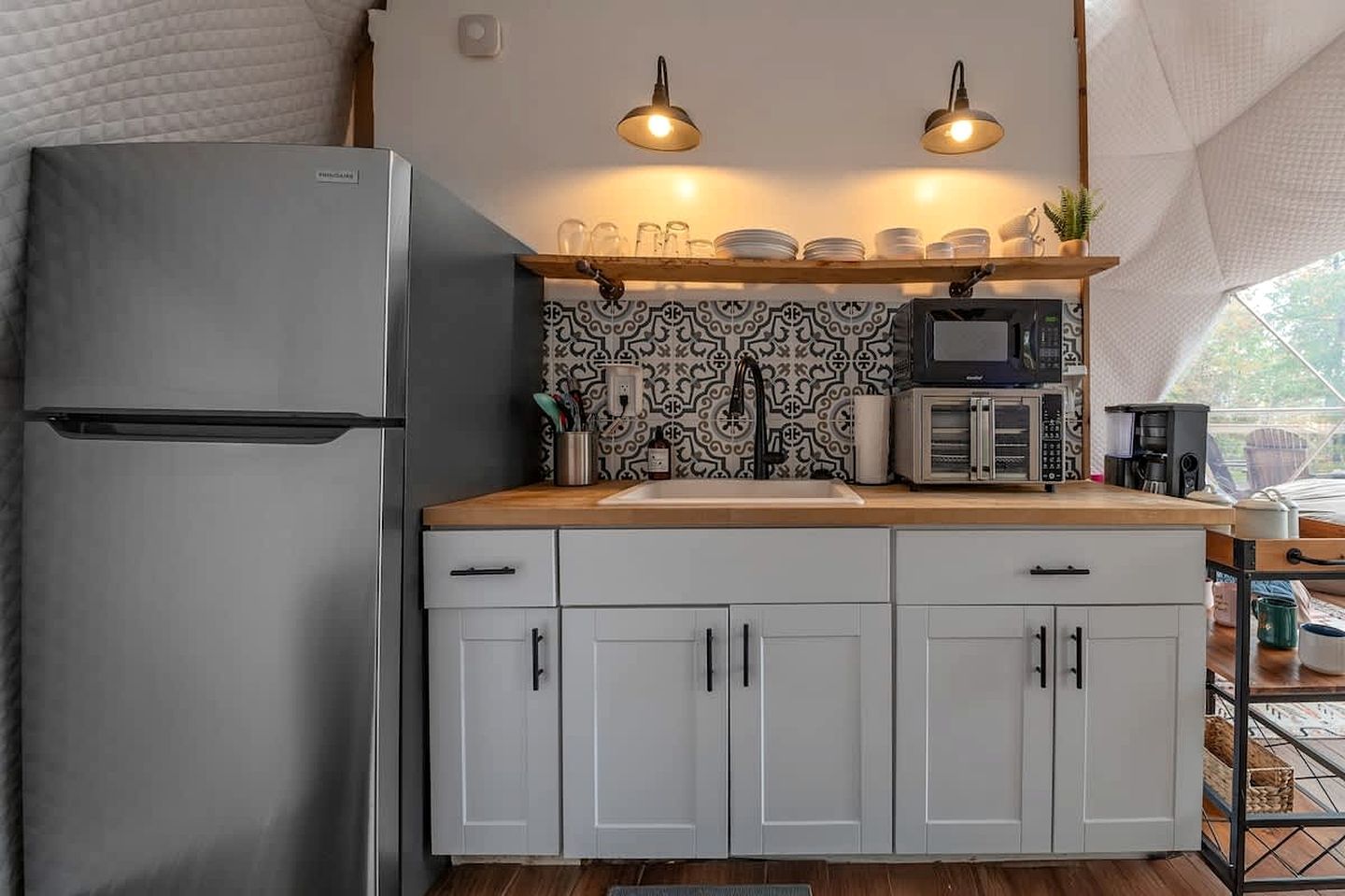A compact kitchen with a refrigerator, microwave, and sink. There are two pendant lights above the counter, and a patterned backsplash.