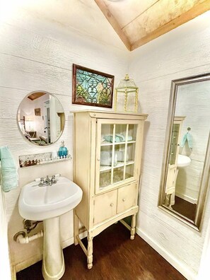 A bathroom with a white sink, a cabinet with glass doors, a round mirror, and a framed picture above the mirror. - Charming Texas Hill Country Cottage near Austin for Romantic Getaways in Texas (Dripping Springs)