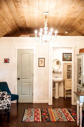 A room with a wooden ceiling, a chandelier, a door, a chair, a sink, a cabinet, and a patterned rug.
