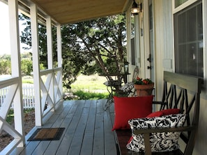A covered porch with a wooden floor, white railings, a bench with patterned cushions, and a view of greenery.