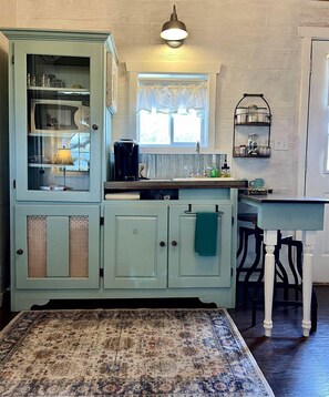 A kitchen with a built-in cabinet, a coffee maker, a sink, and a window with curtains.