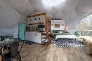 Un interior compacto con forma de tienda de campaña, que incluye una kitchenette, una cama con ropa de cama verde y una zona de comedor con mesa y sillas.