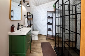 A bathroom with a green vanity, a white toilet, a mirror with two lights, and a black shelving unit with shoes.