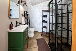 Een badkamer met een groen wastafelmeubel, een wit toilet, een spiegel met twee lampen, en een zwarte schoenenkast.