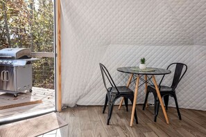 Una habitación con una mesa redonda, dos sillas, una parrilla y vistas a árboles a través de una ventana.