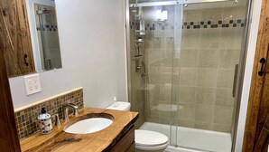 Un baño con tocador de madera, lavamanos blanco, inodoro y cabina de ducha con paredes de vidrio.